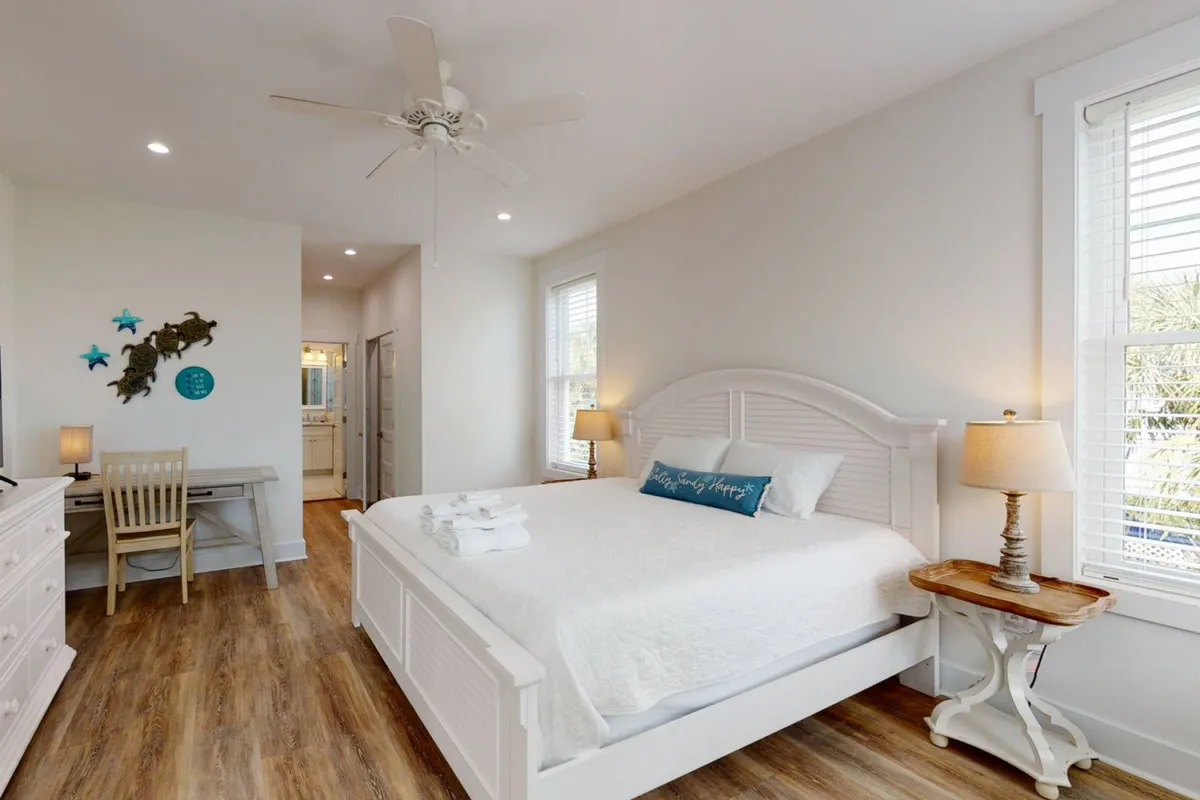 A serene bedroom showcasing a large bed with crisp white linens, a decorative pillow with uplifting text, a wooden side table, and a functional desk with a chair. Soft natural light filters in, creating a calming atmosphere, ideal for unwinding or enjoying a quiet moment.