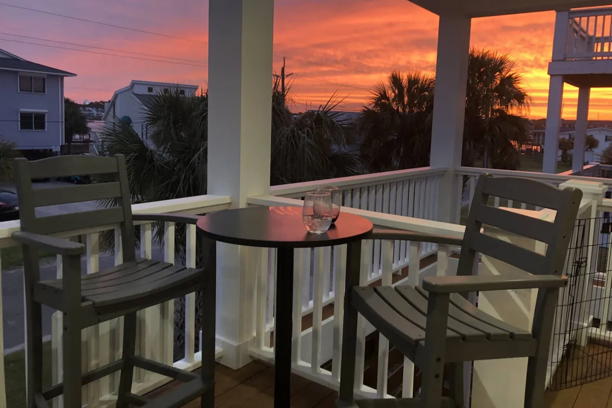 An inviting exterior scene featuring two chairs and a small table facing a brilliant sunset sky, with drinks in hand. The mood is serene, suggesting a peaceful pause after a day of excitement.