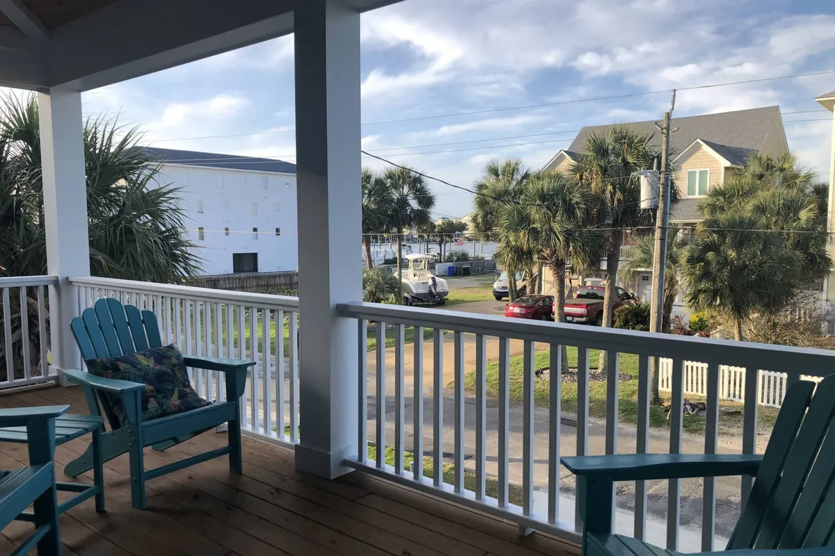 A serene porch scene with Adirondack chairs positioned to overlook a tranquil neighborhood street lined with palm trees and distant houses. The natural light and open air evoke a sense of relaxation and possibility for outdoor work or breaks. The porch area invites guests to step outside, enjoy fresh air, and perhaps take a phone call or sip morning coffee while recharging for the workday.