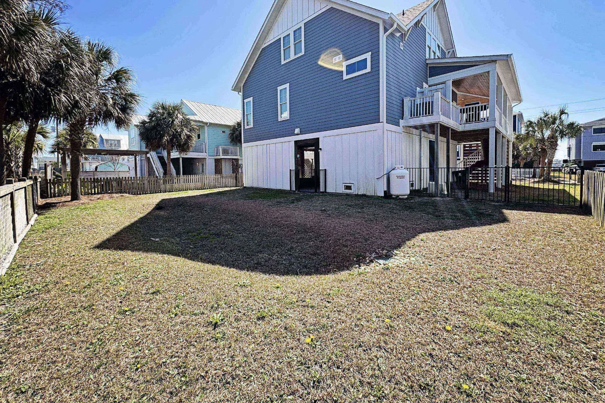 A wide view of the blue house exterior, highlighting the spacious fenced yard with palm trees. The scene is lively, suggesting outdoor gatherings, barbecues, and children playing, all under sunny skies.