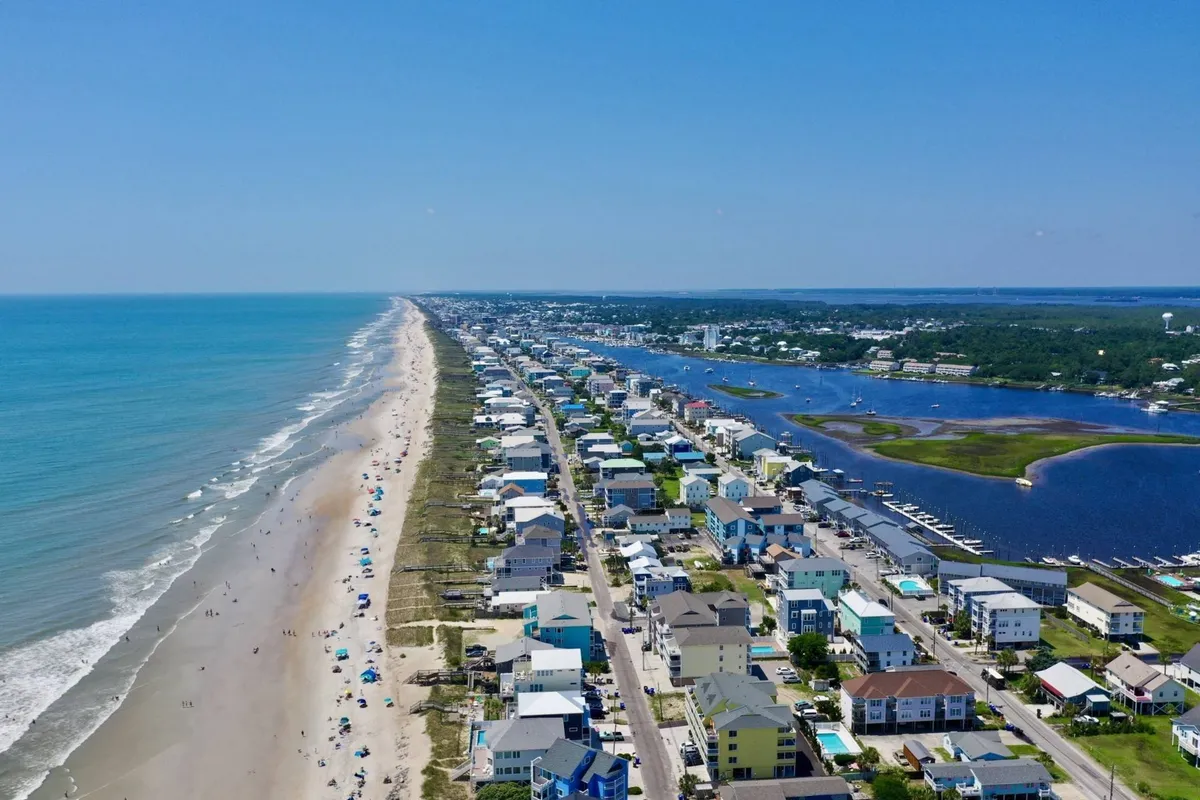 A scenic view of the shoreline with gentle ocean waves, a sandy beach, beach houses, and a marina in the distance. The scene hints at water-based activities and exploration, with visible open water and accessible beachfront.