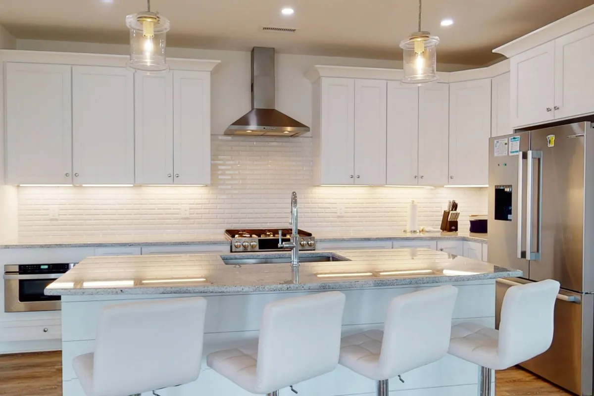 A modern kitchen featuring granite countertops, sleek white cabinetry, stainless steel appliances, and bar stools at a central island. The space is bright, functional, and inviting, ideal for both casual breakfasts and festive meal prep.
