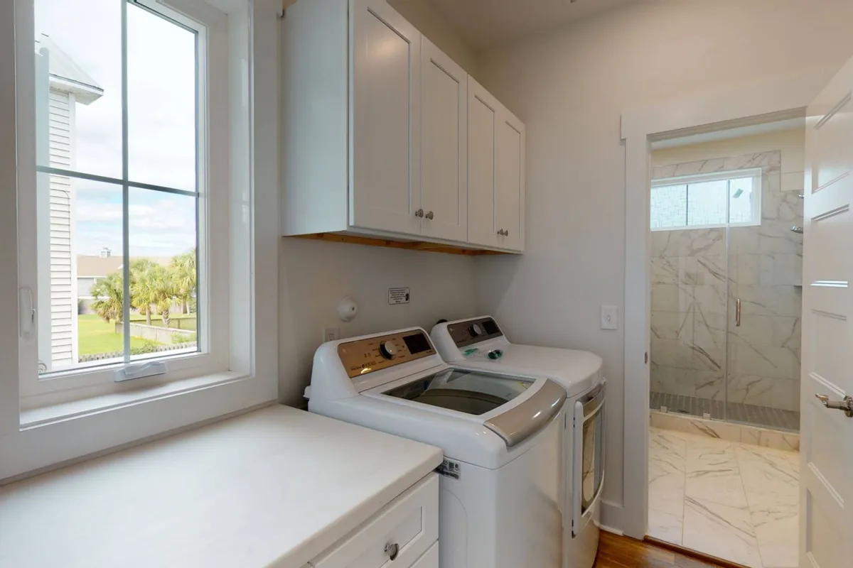 A bright laundry room equipped with a modern washing machine and dryer, a large window providing natural light, and a practical shower area in the background. The space feels both functional and serene, suited for family convenience.