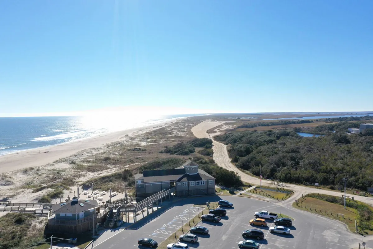 A broad beachfront scene showing the sandy shoreline, parking area, and a coastal building in the backdrop. The wide view suggests immediate proximity to the ocean, evoking the appeal of beach breaks, scenic walks, and after-work leisure. The landscape highlights easy access to outdoor activities and the soothing presence of the coast.