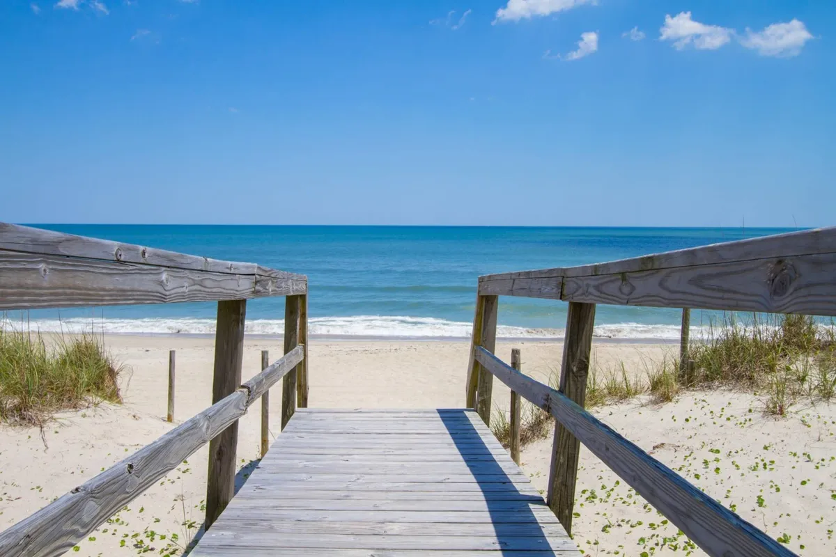 A wooden walkway extends across soft sand toward the blue expanse of the ocean, framed by a bright sky. The scene captures the immediate proximity to the beach, with inviting dunes and gentle waves in the distance. The area feels open and breezy, evoking a sense of easy access to the shore for swimming, sandcastle-building, or relaxing in the sun.