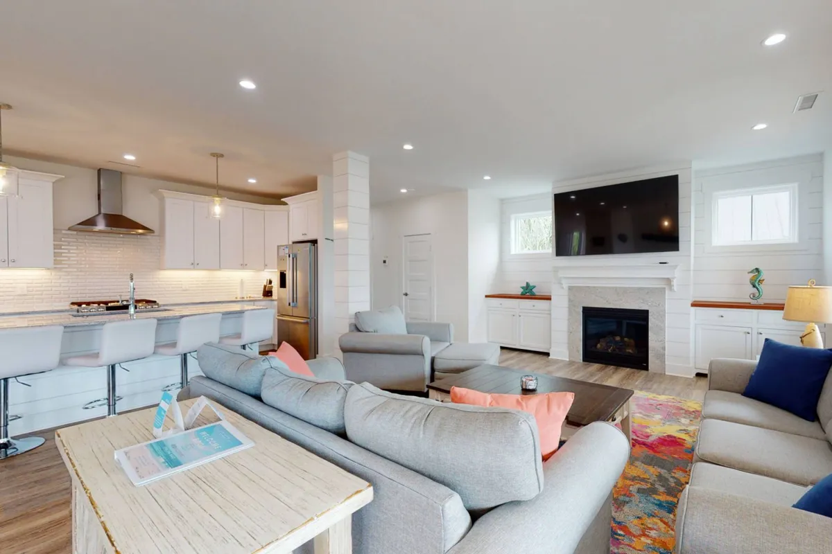 A bright and open living area with a cozy sofa arrangement facing a fireplace and TV, with sightlines extending to a spacious kitchen island. The space exudes warmth and togetherness, perfect for gathering and entertaining.